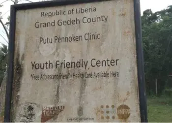 Faded signboard of Putu Pennoken Clinic and Youth Friendly Center in Grand Gedeh County, Liberia, surrounded by overgrown grass and an abandoned building