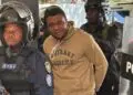 Capitol arson suspect John Nyanti in handcuffs escorted by heavily armed Liberian police officers, including a PSU officer in riot gear, outside the Temple of Justice in Monrovia.