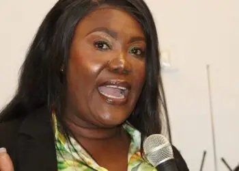 Rochell Woodson speaks into a microphone during a public address, wearing a floral blouse and black blazer.