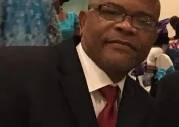 A man wearing a suit and glasses seated indoors during a formal gathering