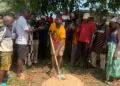 Representative Luther Sandy Collins performs groundbreaking with a shovel as community members gather to launch the maternity waiting home project at Kpayeakwelleh Clinic.