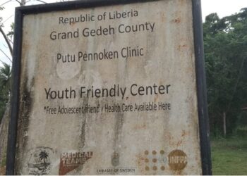 Faded signboard of Putu Pennoken Clinic and Youth Friendly Center in Grand Gedeh County, Liberia, surrounded by overgrown grass and an abandoned building