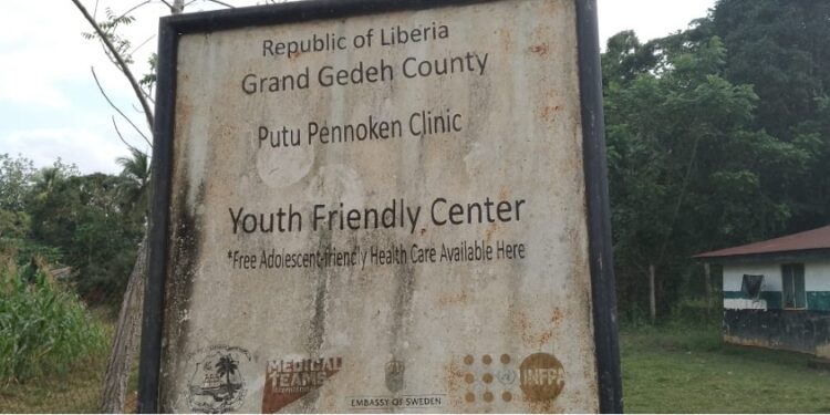 Faded signboard of Putu Pennoken Clinic and Youth Friendly Center in Grand Gedeh County, Liberia, surrounded by overgrown grass and an abandoned building