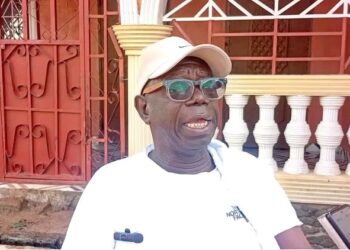 Rep. Robert Flomo Womba wearing a white cap and shirt, speaking to journalists outside his residence in Bong County about the Bong University bill.
