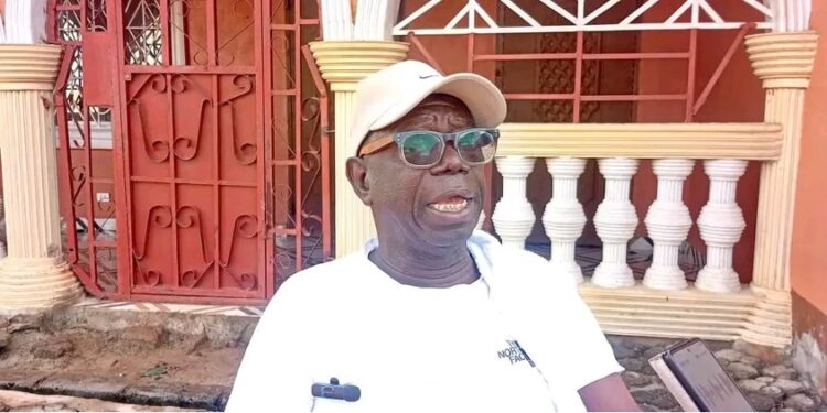 Rep. Robert Flomo Womba wearing a white cap and shirt, speaking to journalists outside his residence in Bong County about the Bong University bill.