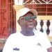 Rep. Robert Flomo Womba wearing a white cap and shirt, speaking to journalists outside his residence in Bong County about the Bong University bill.