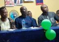 peaker Richard Koon (center) seated at a blue-draped table during a community outreach event in West Point, Monrovia, flanked by Representative Emmanuel Dahn and local leaders, with green balloons and political posters visible in the background