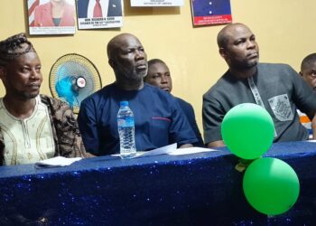 peaker Richard Koon (center) seated at a blue-draped table during a community outreach event in West Point, Monrovia, flanked by Representative Emmanuel Dahn and local leaders, with green balloons and political posters visible in the background