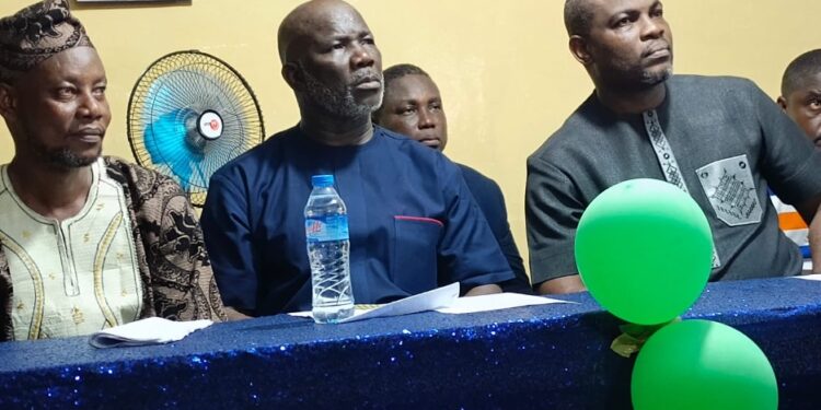 peaker Richard Koon (center) seated at a blue-draped table during a community outreach event in West Point, Monrovia, flanked by Representative Emmanuel Dahn and local leaders, with green balloons and political posters visible in the background