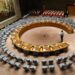 A wide-angle view of the United Nations Security Council chamber with empty seats arranged in a circular formation, symbolizing global diplomacy and decision-making.