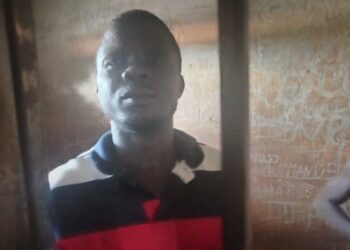 A man wearing a red, black, and white striped shirt stands behind metal bars in a dimly lit detention cell, with graffiti visible on the wall behind him