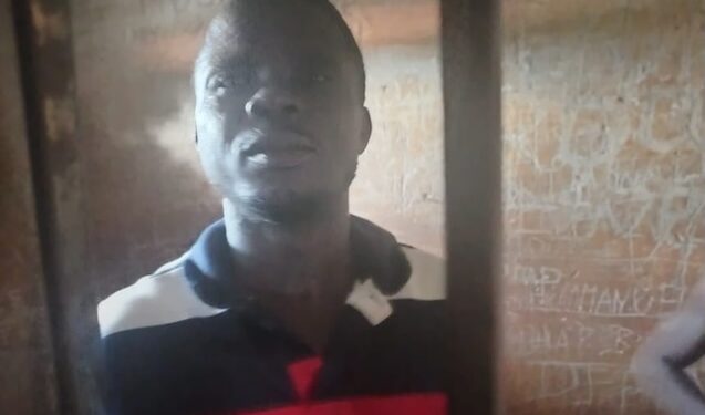 A man wearing a red, black, and white striped shirt stands behind metal bars in a dimly lit detention cell, with graffiti visible on the wall behind him