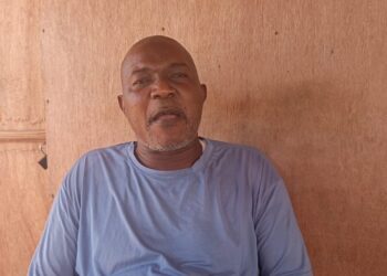 A middle-aged man wearing a light blue shirt sits against a wooden wall, speaking directly to the camera. He appears serious and composed, possibly giving a statement or interview.