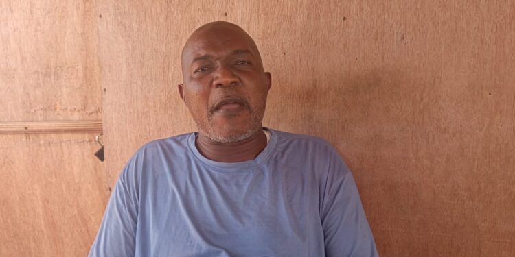 A middle-aged man wearing a light blue shirt sits against a wooden wall, speaking directly to the camera. He appears serious and composed, possibly giving a statement or interview.