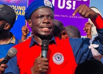 CDC Youth League Chairman Emmanuel M. Johnson raises his fist while speaking into a microphone during a protest, with a “CDC Lives in Us” banner visible in the background