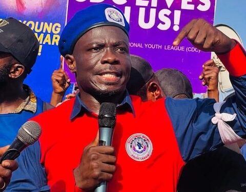 CDC Youth League Chairman Emmanuel M. Johnson raises his fist while speaking into a microphone during a protest, with a “CDC Lives in Us” banner visible in the background