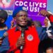 CDC Youth League Chairman Emmanuel M. Johnson raises his fist while speaking into a microphone during a protest, with a “CDC Lives in Us” banner visible in the background