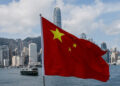 Chinese national flag waves prominently in front of the Hong Kong skyline, with Victoria Harbour and skyscrapers including the International Finance Centre visible in the background under a partly cloudy sky