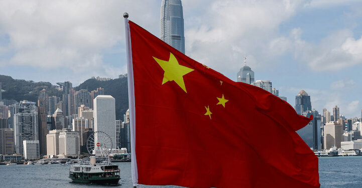 Chinese national flag waves prominently in front of the Hong Kong skyline, with Victoria Harbour and skyscrapers including the International Finance Centre visible in the background under a partly cloudy sky