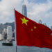 Chinese national flag waves prominently in front of the Hong Kong skyline, with Victoria Harbour and skyscrapers including the International Finance Centre visible in the background under a partly cloudy sky