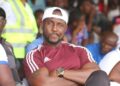Former Liberia international James Debbah sits at a football match wearing a maroon Adidas shirt and white cap, now facing backlash over Ligue 1 vs Premier League remarks.
