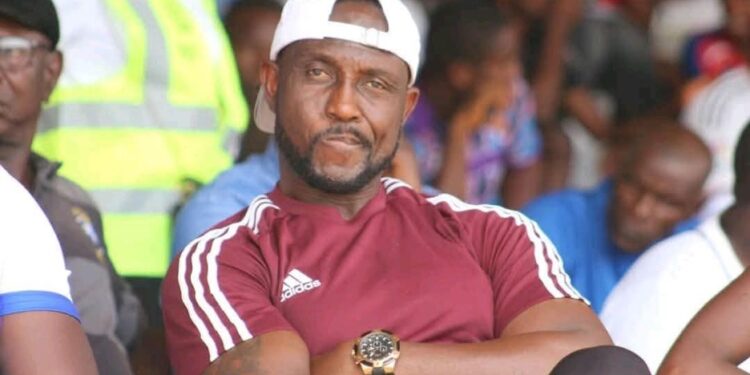 Former Liberia international James Debbah sits at a football match wearing a maroon Adidas shirt and white cap, now facing backlash over Ligue 1 vs Premier League remarks.