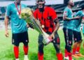Liberian footballer Jegbay Morris Konneh celebrates with a trophy and teammates on the field, wearing a red and black jersey after a tournament win.