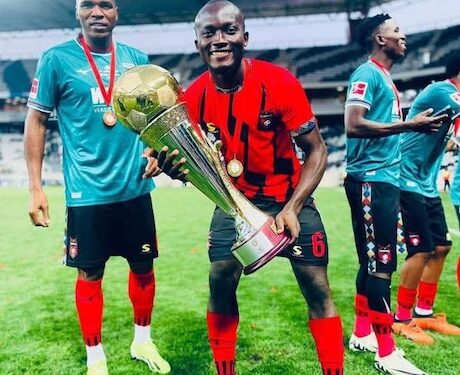 Liberian footballer Jegbay Morris Konneh celebrates with a trophy and teammates on the field, wearing a red and black jersey after a tournament win.