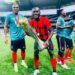 Liberian footballer Jegbay Morris Konneh celebrates with a trophy and teammates on the field, wearing a red and black jersey after a tournament win.