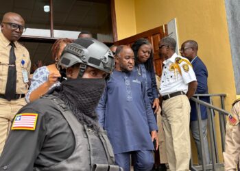 Ambassador Dee-Maxwell Saah Kemayah exits a courthouse in Monrovia surrounded by security personnel, including an armed officer in tactical gear and uniformed court officers, following his arrest in connection with a corruption investigation.