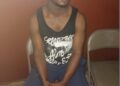 Lawrence K. Williams, suspect in the alleged vehicular attack on Unity Party supporters, seated in a holding area wearing a black sleeveless shirt and blue shorts, hands clasped, awaiting trial amid stalled proceedings