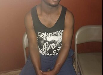 Lawrence K. Williams, suspect in the alleged vehicular attack on Unity Party supporters, seated in a holding area wearing a black sleeveless shirt and blue shorts, hands clasped, awaiting trial amid stalled proceedings