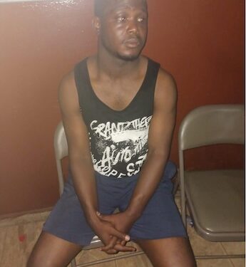 Lawrence K. Williams, suspect in the alleged vehicular attack on Unity Party supporters, seated in a holding area wearing a black sleeveless shirt and blue shorts, hands clasped, awaiting trial amid stalled proceedings