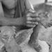 A man affected by leprosy sits barefoot on a concrete platform at Suakoko Rehabilitation Center, Bong County, Liberia, showing damaged limbs and torn clothing.