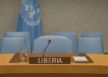 Liberia’s designated seat at the United Nations Security Council, featuring a wooden desk with a nameplate labeled “LIBERIA,” a microphone, a glass of water, documents, and several light blue chairs in the background, with the UN flag hanging behind.
