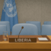 Liberia’s designated seat at the United Nations Security Council, featuring a wooden desk with a nameplate labeled “LIBERIA,” a microphone, a glass of water, documents, and several light blue chairs in the background, with the UN flag hanging behind.