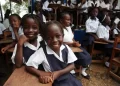 Smiling Liberian schoolgirls in uniform sit at wooden desks in an outdoor classroom setting, surrounded by classmates, during a school session.