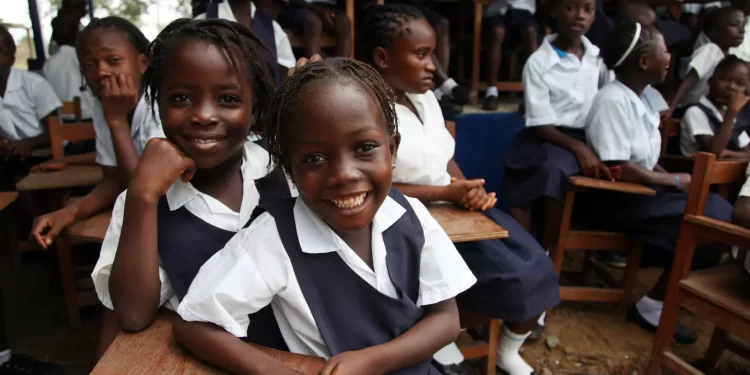 Smiling Liberian schoolgirls in uniform sit at wooden desks in an outdoor classroom setting, surrounded by classmates, during a school session.
