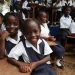 Smiling Liberian schoolgirls in uniform sit at wooden desks in an outdoor classroom setting, surrounded by classmates, during a school session.
