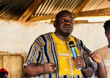 Musa Bility speaks passionately while holding a microphone at a political gathering in Kakata, wearing a traditional yellow and striped Liberian shirt, as supporters listen attentively in the background
