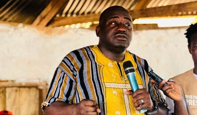 Musa Bility speaks passionately while holding a microphone at a political gathering in Kakata, wearing a traditional yellow and striped Liberian shirt, as supporters listen attentively in the background