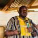 Musa Bility speaks passionately while holding a microphone at a political gathering in Kakata, wearing a traditional yellow and striped Liberian shirt, as supporters listen attentively in the background
