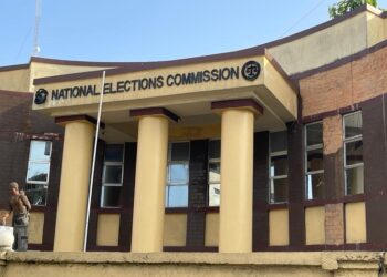Front view of the National Elections Commission (NEC) headquarters building in Monrovia, Liberia, with yellow columns and maroon trim, and a man standing near the entrance.