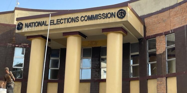 Front view of the National Elections Commission (NEC) headquarters building in Monrovia, Liberia, with yellow columns and maroon trim, and a man standing near the entrance.