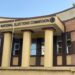 Front view of the National Elections Commission (NEC) headquarters building in Monrovia, Liberia, with yellow columns and maroon trim, and a man standing near the entrance.