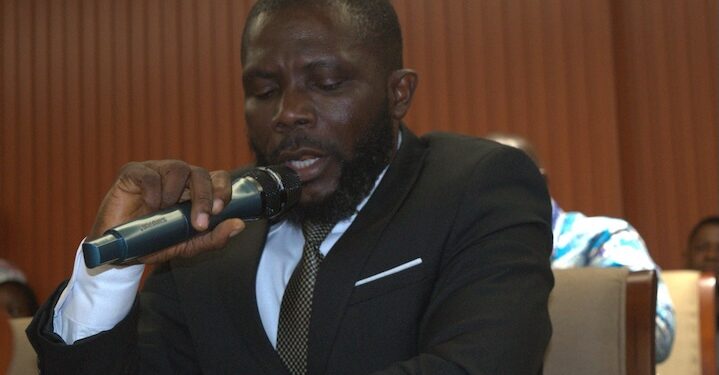 Rep. Jacob Debee speaks into a microphone during a legislative session, wearing a dark suit and patterned tie.