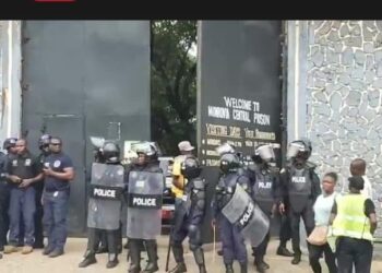 Riot police stand guard at the entrance of Monrovia Central Prison as a group of officers and civilians look on during the detention of lawmakers accused in the Capitol Building arson case