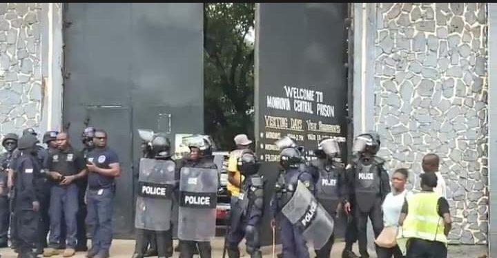 Riot police stand guard at the entrance of Monrovia Central Prison as a group of officers and civilians look on during the detention of lawmakers accused in the Capitol Building arson case