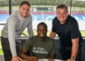Christian Saydee, wearing a Wigan Athletic shirt, poses with two club officials after signing a three-year contract at the Brick Community Stadium ahead of the 2025–26 League One season.