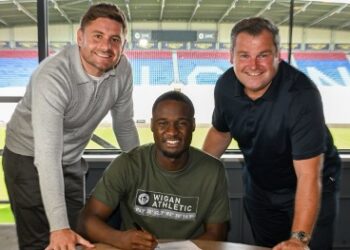 Christian Saydee, wearing a Wigan Athletic shirt, poses with two club officials after signing a three-year contract at the Brick Community Stadium ahead of the 2025–26 League One season.